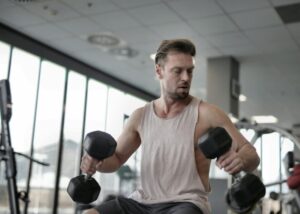 From below of muscular male pumping biceps with dumbbells while sitting on bench in sports center